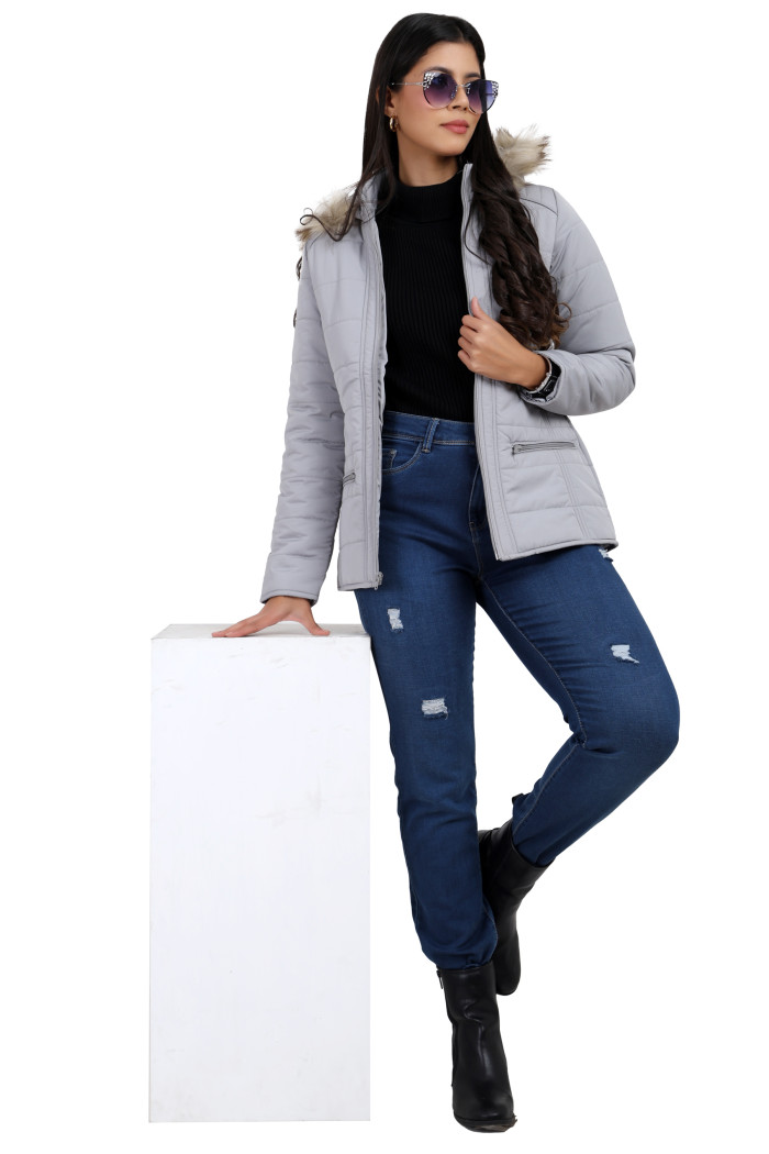 A woman in a standing pose in black shades, wearing Trufit’s grey Matte Quilted Jacket with Horizontal Pockets, zip closure, a removable hood and blue jeans, is holding the jacket with her left hand.